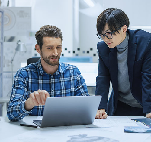 Modern Factory: Industrial Engineer Sitting at His Desk, Working on Laptop Computer, Talks with Chief Project Manager, They Analyze Mechanisms and Blueprints. Manufactory with CNC Working Machinery